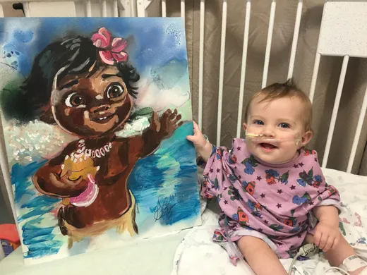 A baby smiling beside a hand-painted Moana painting
